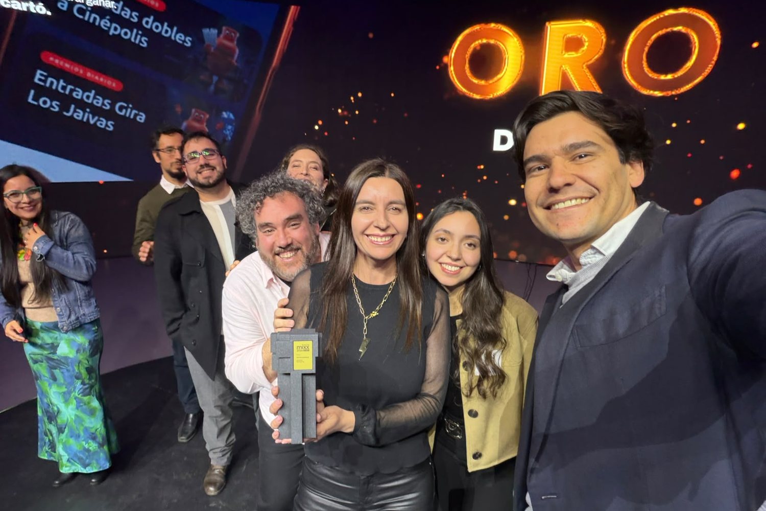 Siete personas sonríen en el escenario de los IAB MIXX; una sostiene el trofeo y detrás, en letras naranjas gigantes, dice ORO.