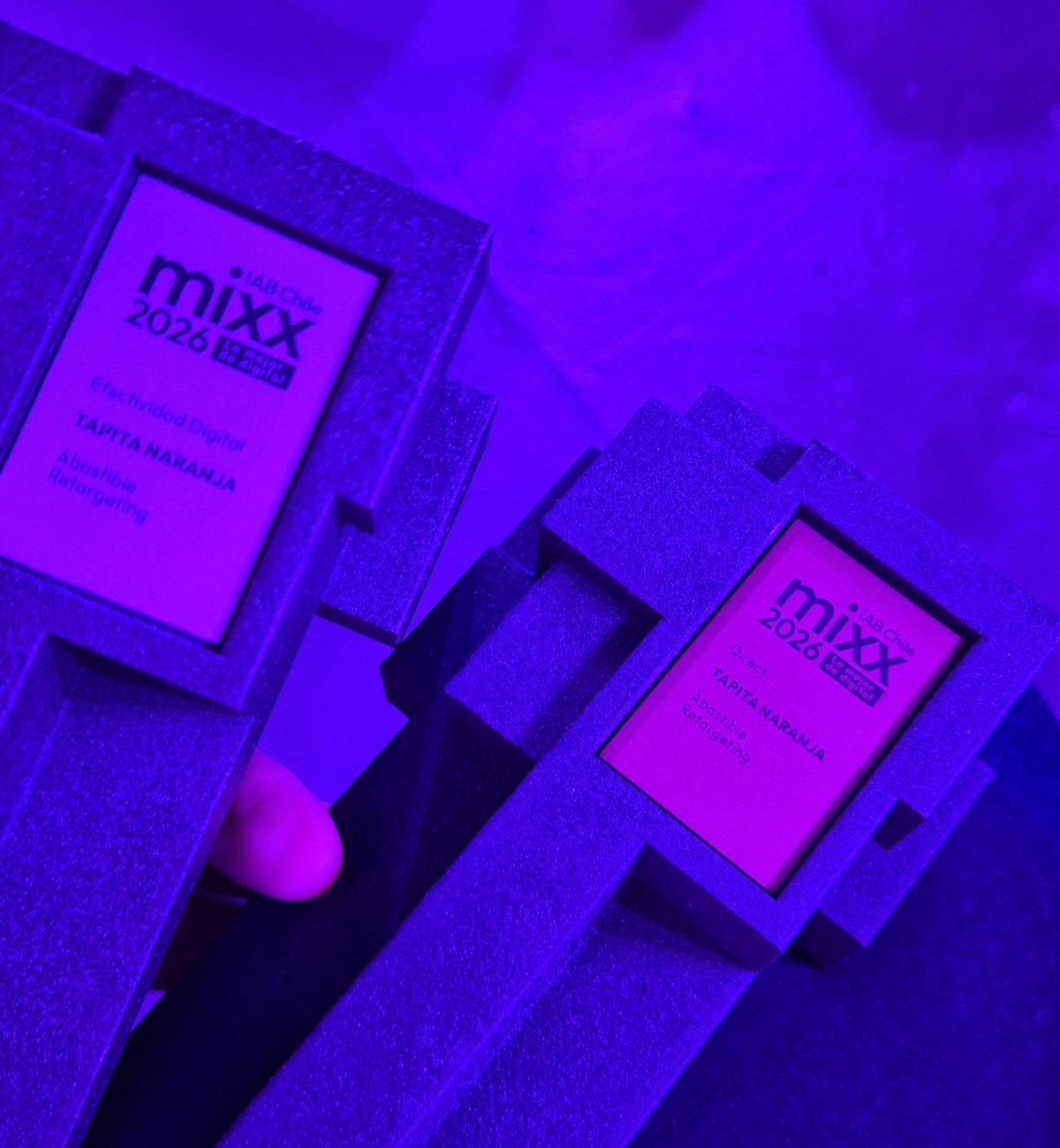 Los dos trofeos de IAB MIXX 2026, uno al lado del otro, bajo la luz violeta de la fiesta: Efectividad Digital y Directo, Tapita Naranja, Abastible, Retargeting.